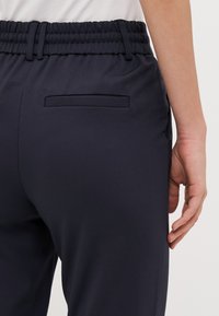 Marineblå joggebukser med en rynket elastisk linning, som har en liten, lomme på baksiden og en jevn stofftekstur.