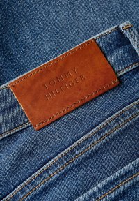 Vaqueros de mezclilla de tonalidad azul, con una parche de cuero marrón etiquetado como "TOMMY HILFIGER". Costuras visibles en un color contrastante.