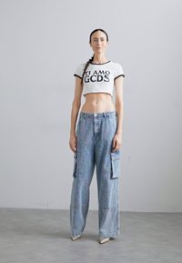 T-shirt branca curta com letras pretas, combinada com calças cargo largas azul-claro com bolsos frontais e brilho metálico.