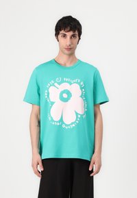 Turquoise cotton t-shirt featuring a large pink flower graphic and circular text design. Short sleeves and a relaxed fit.