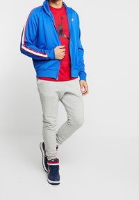 Chaqueta de chándal azul con cremallera y rayas rojas y blancas, combinada con una camiseta roja con un gráfico negro y pantalones de chándal grises.