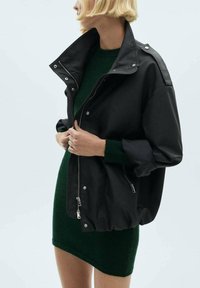 Giacca nera, oversize, con colletto alto, chiusura con zip e bottoni a pressione, e tasche laterali, indossata sopra un vestito in maglia verde. Tessuto liscio, design casual.
