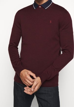 Mann trägt einen burgunderfarbenen Pullover über einem marineblauen Hemd mit Kragen und gestreiftem Besatz, steht mit vor der Brust verschränkten Händen, dunkle Hose sichtbar.