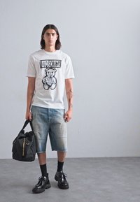 Tânăr îmbrăcat cu un tricou alb cu ursuleț Moschino, pantaloni scurți din denim distruși, șosete și pantofi negri, ținând o geantă neagră în fața unui zid gri simplu.