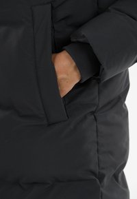 Schwarze gepolsterte Jacke mit einer Seitentasche und gerippten Bündchen. Der Stoff ist glatt und matt, was Wärme und Funktionalität betont.