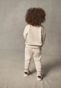 Kermanvärinen collegepaita ja siihen sopivat housut, tehty pehmeästä kankaasta. Takaa näkyy kiharat hiukset. Tennarit, joissa mustat yksityiskohdat.