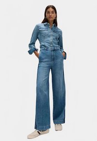 Džínový overal světle modré barvy, s dlouhými rukávy, zapínáním na knoflíky vpředu, dvěma náprsnými kapsami, širokými nohavicemi a manžetovými lemy.