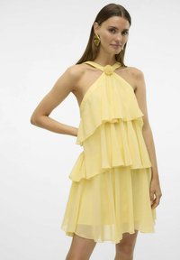Ærmeløs gul chiffon kjole med lagdelte volanger og en knudedetail ved halsen. Glat overflade med en svævende silhuet.