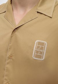 Gros plan d’une chemise beige à boutons avec des coutures blanches et un patch logo brodé sur la poitrine représentant un court de tennis et un crocodile.