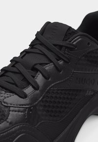 Gros plan sur une basket noire avec des panneaux texturés et lisses, des lacets noirs et le logo "HUGO" sur le côté.