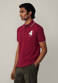 Bordový polo tričko s bielym lemovaním a golierom. Na prednej strane je biele číslo "4" a názov značky "HACKETT". Krátke rukávy.