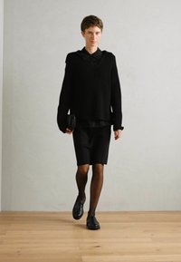 Crna pletena majica s kapuljačom, uska crna suknja i prozirne crne tajice. Model nosi crne cipele i drži mali clutch.