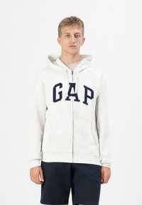 Hvit zip-up hettegenser med en stor marineblå "GAP" logo, frontlommer og snor i hetten. Brukt med marineblå shorts.