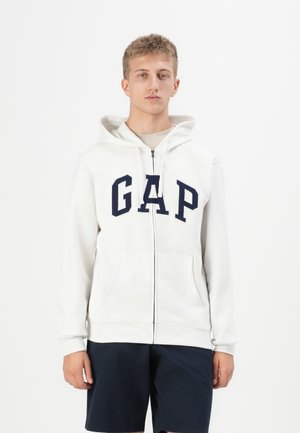 White zip-up hoodie featuring a large navy "GAP" logo, front pockets, and a drawstring hood. Worn with navy shorts.