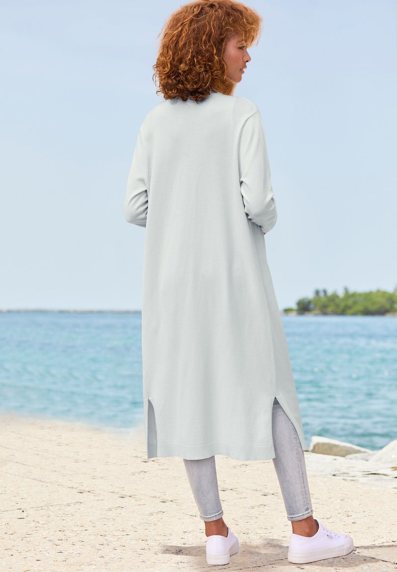 Cardigan lungo grigio chiaro con design dritto, fessure laterali e maniche lunghe, abbinato a jeans chiari e sneakers bianche.