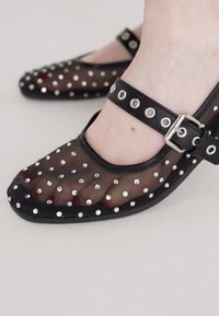 Chaussures noires transparentes ornées de clous argentés, dotées d'un bout arrondi et d'une bride de cheville avec une boucle. Le matériau texturé ajoute un intérêt visuel.