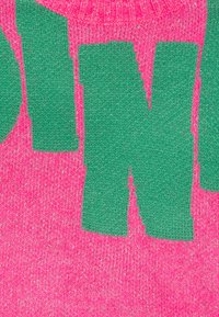 Pull en tricot avec des lettres vertes audacieuses sur un fond rose vif. La texture est douce et le design comprend une finition pailletée.