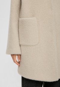 Beige wollen blendjas met een gestructureerde afwerking, voorzien van een patchzak en brede mouwen, getoond tot kort bij de arm en heup.