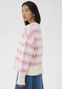 Jeune femme aux longs cheveux bruns portant un pull blanc à rayures roses et une jupe en denim bleu, de profil, sur un fond uni.