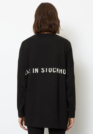 Person med skulderlangt brunt hår iført en sort sweatshirt med lange ærmer, hvor hvid tekst "STOCKHO" delvis er synlig på ryggen.