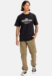 Camiseta negra de algodón con el logo blanco de Quiksilver; pantalones cargo color caqui con bolsillos laterales; zapatillas negras con suelas blancas.