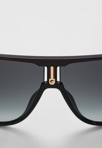 Ochelari de soare negri cu lentile gri degradé, având un accent auriu proeminent pe punte și un design geometric îndrăzneț în montură.