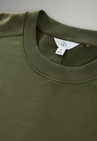 Olive rohelise värvi ümar kaelusega sügisjakk, ribidega kaeluse ja varrukaotstega. Etikett kannab nime "THE SET," suurus M, valmistatud Bangladeshi. Pehme tekstuur, minimaalne disain.