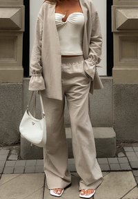 Kvinna klädd i beige lösa byxor och blazer, vit rynkad strapless topp, vita sandaler, hållande en vit handväska, stående på stenbeläggning.