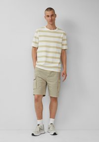 Weißes Kurzarmhemd mit gelben Streifen, kombiniert mit hellbeigen Cargo-Shorts. Graue Sportschuhe mit weißen Socken. Neutraler Hintergrund.