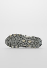 Semelle d’une chaussure de sport grise et beige avec des motifs de semelle profonds pour l’adhérence, montrée sur un fond blanc uni.