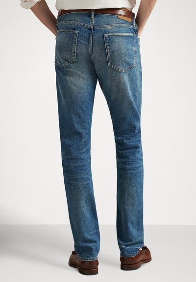 Vīrietis, kurš valkā zilas džinsa bikses, brūnu ādas jostu, baltu kreklu un brūnas ādas klasiskās kurpes, stāvot ar rokām kabatās, skats no muguras.