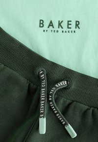 Zaļas sporta bikses ar piparmētru zaļu josta un auklu. Uz auduma ir melnā krāsā redzams zīmola nosaukums "BAKER by TED BAKER". Gluda tekstūra.