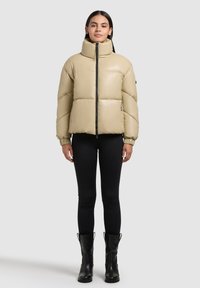 Beige színű, magas nyakú puffermellény, elülső cipzárral és elasztikus mandzsettával. Fekete leggings-szel és térd fölé érő fekete csizmával párosítva. Sima textúra.