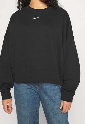 Personne portant un sweat-shirt noir Nike avec un logo blanc sur la poitrine et un jean en denim bleu, coupé à la taille.