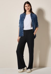 Blauwe denim overhemd over een wit t-shirt, gecombineerd met zwarte elastische broek en witte sneakers met zwarte accenten. Eenvoudig ontwerp, casual pasvorm.