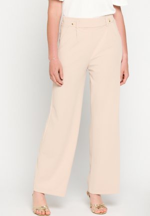 Nainen, jolla on yllään beiget leveälahkeiset housut kultaisilla nappiyksityiskohdilla ja kultaiset avoimet korkokengät, seisoo yksinkertaista vaaleaa taustaa vasten.