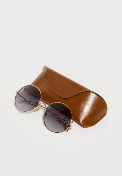 Στρογγυλά γυαλιά ηλίου με χρυσό πλαίσιο και σκοτεινούς φακούς με διαβάθμιση, δίπλα σε καφέ δερμάτινη θήκη με κλείσιμο με πτερύγιο, σε λευκό φόντο.