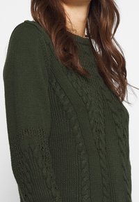 Suéter de punto verde oscuro con patrones de trenzado, mangas texturizadas y cuello de canalé. Tejido suave de peso medio.
