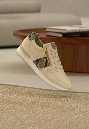 Bézs sportcipő texturált, kígyómintás díszítéssel az oldalakon, fűzős kialakítással, arany cipzár részlettel és fehér gumitalppal.