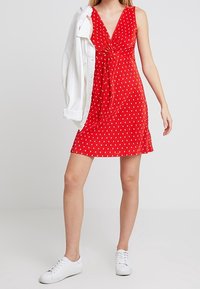 Robe sans manches rouge avec un devant torsadé, des pois blancs et un ourlet fluide. Associée à une veste en denim blanche et des baskets blanches.