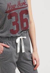Tuta grigia senza maniche con grafica "New York 36" in bordeaux. Presenta una fascia elastica in vita con cordino bianco e tasche laterali. Tessuto morbido.