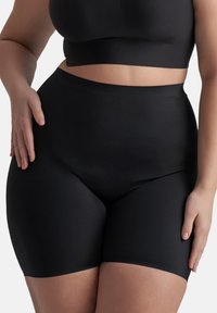 Donna che indossa dei pantaloni a vita alta senza cuciture neri e un top corto nero, con le mani appoggiate sui fianchi e le cosce in vista.
