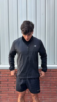 Man die een zwart vest met ritssluiting en New Balance-logo aanpast, zwarte shorts draagt, staand tegen een muur van baksteen en metaalpanelen.