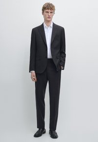 Completo scuro su misura con rever a incavo, abbinato a una camicia bianca con bottoni. Scarpe in pelle nera completano l'outfit. Design minimalista.