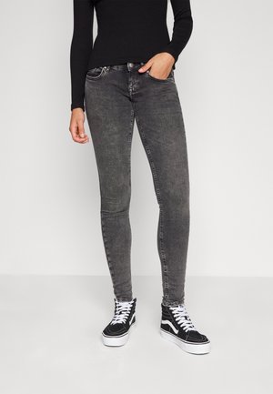 Person wearing high-waisted faded black skinny jeans, black long-sleeve top, and black-and-white high-top sneakers, standing against white background.