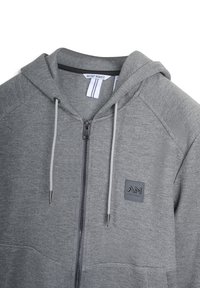 Sudadera con cremallera gris hecha de tejido suave. Cuenta con capucha, cordones y un parche con el logo. Incluye una cremallera de longitud completa y bolsillos frontales.