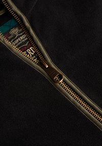 Czarna tkanina polarowa z oliwkowym zamkiem błyskawicznym i złotymi elementami, posiadająca teksturowaną metkę wewnętrzną z detalami marki.