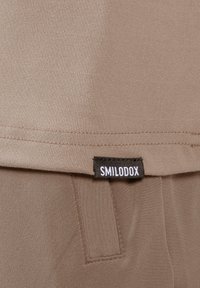 Beiger Sportstoff mit glatter Textur, versehen mit einem gesteppten Saum und einem schwarzen Etikett mit der Aufschrift "SMILODOX" am unteren Rand.