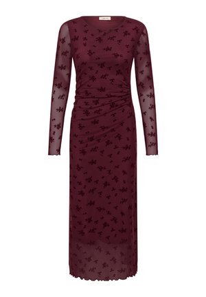 Une femme porte une robe midi bordeaux à imprimé floral et à manches longues, ainsi que des sandales, sur un fond clair et uni.