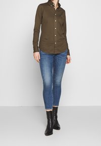 Hnědá košile s límečkem, dlouhými rukávy a slim fit, doplněná modrými skinny džíny a černými kotníkovými botami.